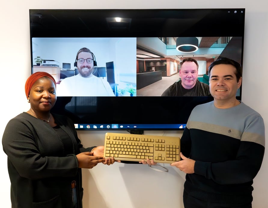 Quatre personnes : deux en personne debout devant un écran; deux à l’écran. Les deux personnes présentes tiennent un trophée doré en forme de clavier.