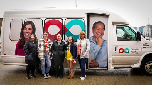 Cinq membres du personnel de Payworks lors d’une journée nuageuse de printemps se tenant devant un autobus sur lequel il est écrit Société canadienne du sang.