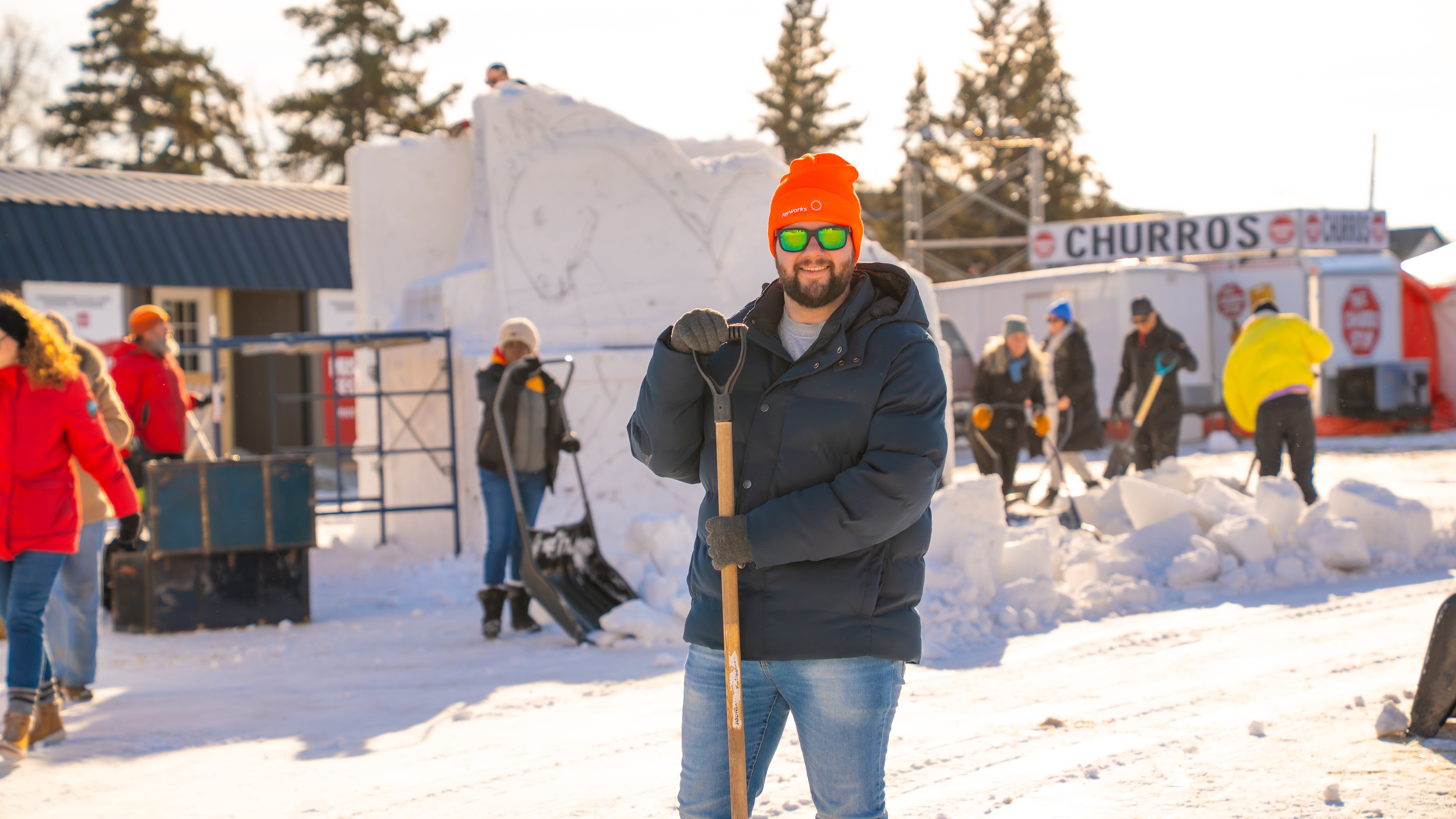 A person wearing an orange Payworks toque and holding a shovel stands in a busy outdoor space, with several people in the background moving snow.