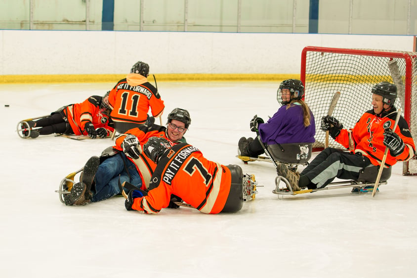 Six people on ice in sledges, almost all wearing orange Payworks jerseys. Several people are toppled over.