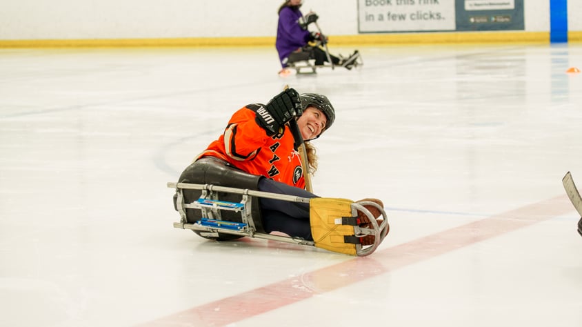 One smiling person wearing an orange Payworks jersey is tipped over in a sledge.