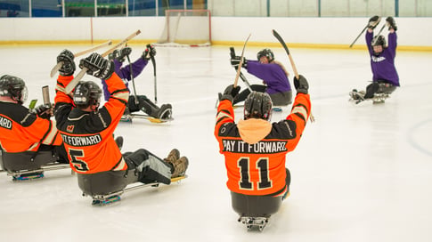Un groupe de personnes en chandail orange de Payworks devant un groupe de personnes en chandail violet de Manitoba Possible qui leur apprennent comment se propulser en hockey sur luge.