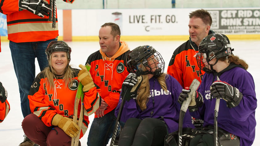 A group of people wearing orange Payworks jerseys and purple Manitoba Possible jerseys sitting on-ice in an arena and smiling together.