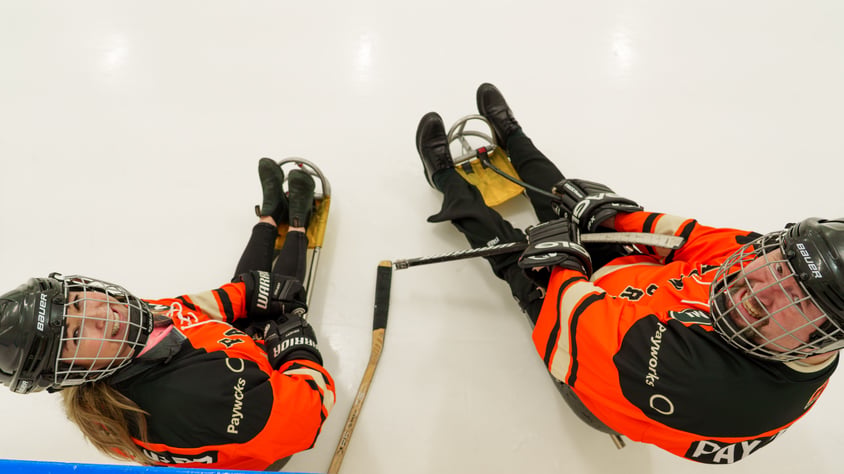 Two people in orange Payworks jerseys sitting on ice and smiling up at the camera.