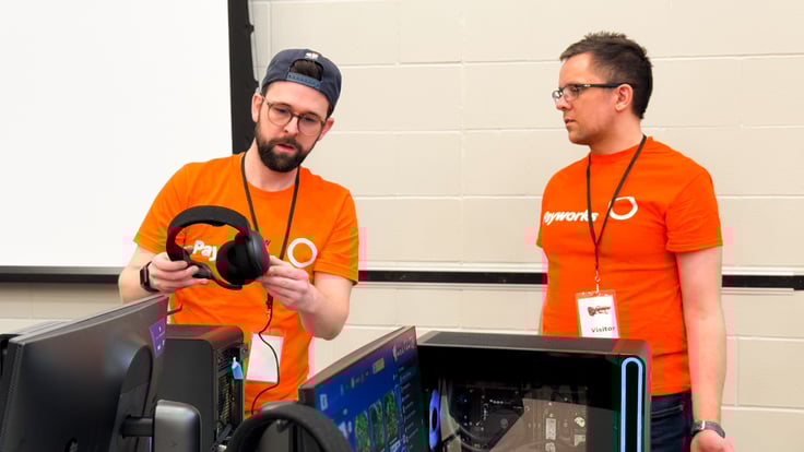 Deux hommes en chandail Payworks se tiennent devant des écrans d’ordinateur; l’un deux tient un casque d’écoute.