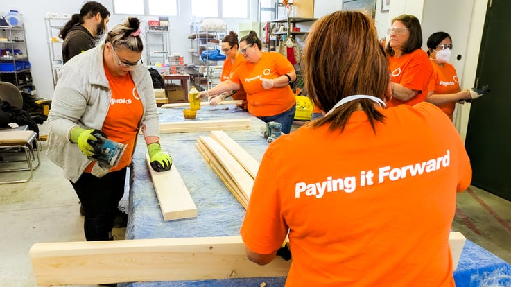 Un groupe de personnes en chandail orange « Donner au suivant » ponce des lattes en bois.