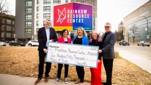 Five people holding an oversized cheque in front of an exterior sign that reads Rainbow Resource Centre.
