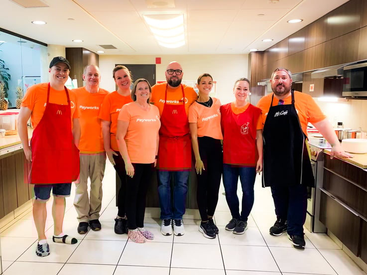 Huit personnes en chandail orange Payworks sourient pour la photo dans une cuisine commerciale.