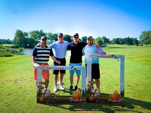 Quatre membres du personnel de Payworks sur un terrain de golf, devant un panneau portant l’inscription « J’aime les camps Tims. »