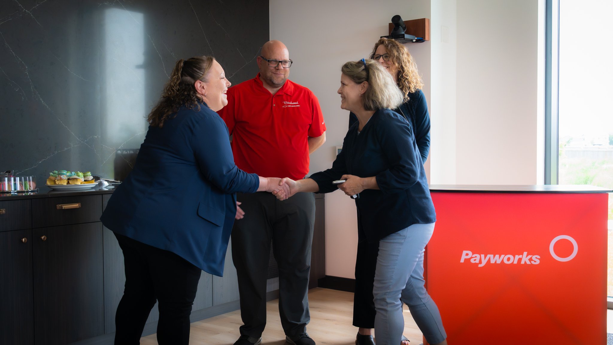 Two people shake hands and smile in front of a Payworks branded backdrop, while two others look on. A spread of cupcakes is visible on a counter in the background.