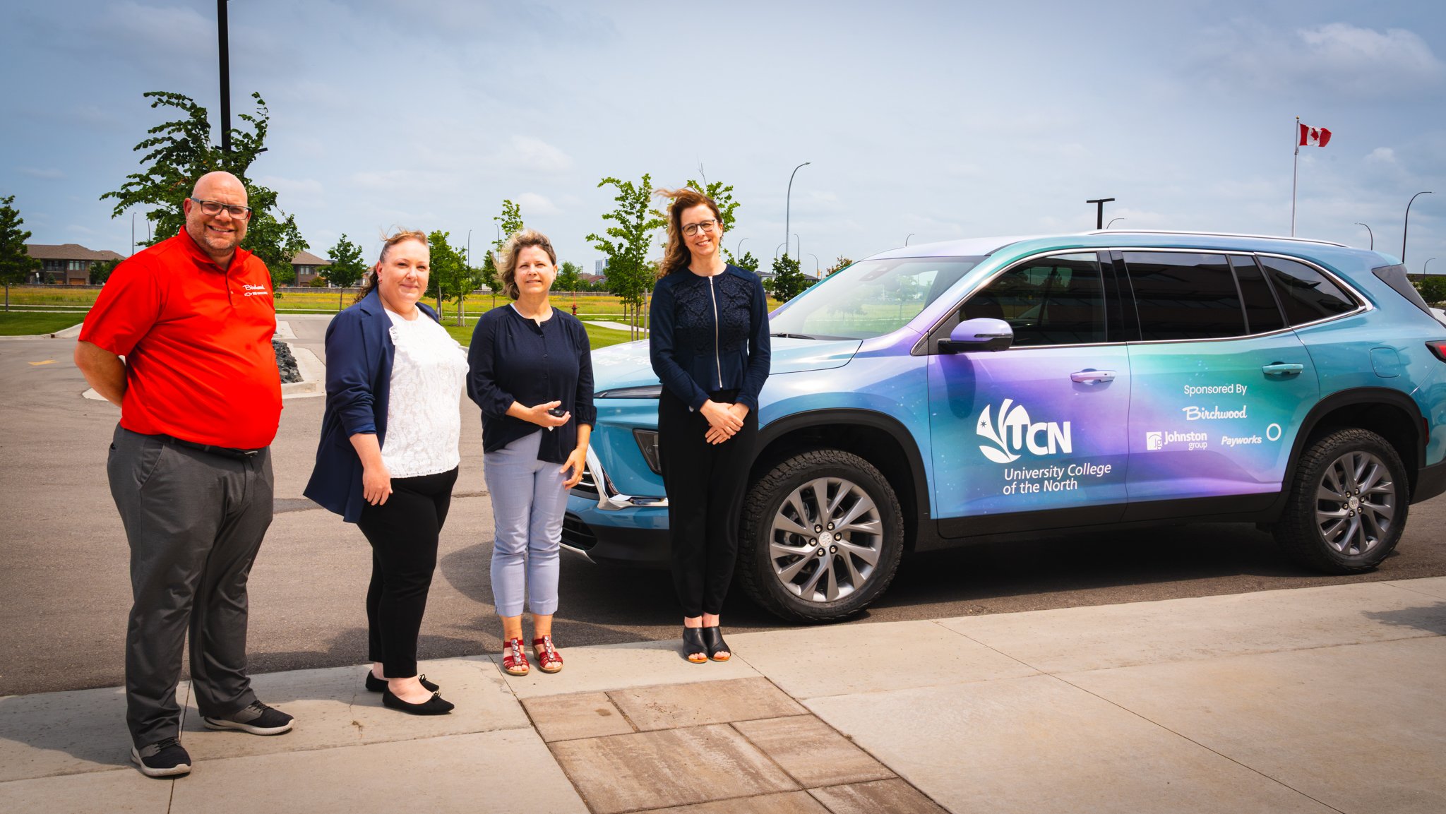 Four people stand outside in a parking lot, beside a vehicle that is coloured in a wrap of the Northern Lights, with company logos on it of "University College of the North", "Birchwood", "Johnston Group" and "Payworks". Trees blow in the wind behind them.
