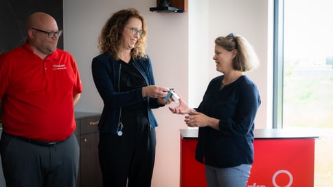 Trois personnes se tiennent ensemble devant un podium rouge. La personne au centre remet un jeu de clés à la personne de droite, et elles sourient toutes les deux.