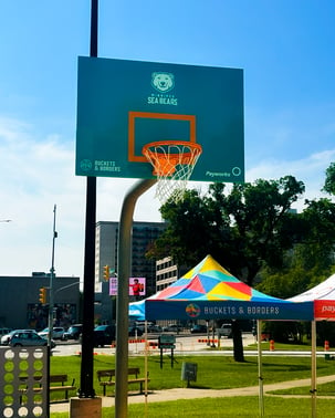 Un panier de basket-ball au Mayfair Project ; il est bleu sarcelle et porte les logos des Sea Bears, Buckets & Borders et Payworks.