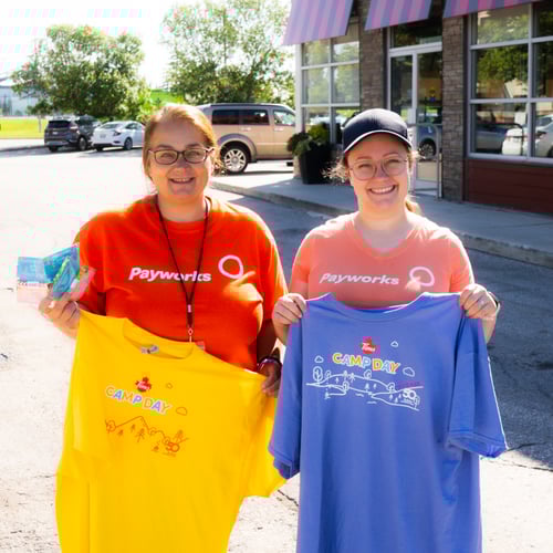 Two Payworks staff in matching orange t-shirts holding two Camp Day t-shirts.