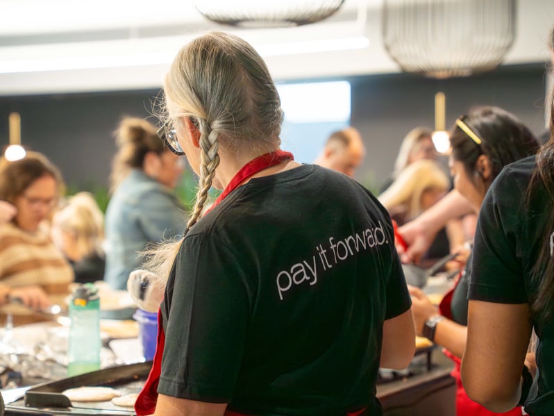 Une personne qui porte un chandail sur lequel on peut lire « pay it forward » et un tablier rouge prépare des crêpes pour un groupe de personnes.