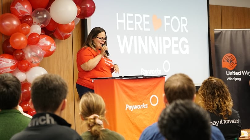 A United Way Sponsored Campaign Associate addressing a crowd through a microphone behind a Payworks-branded podium and beside a sign that reads “Here for Winnipeg.”