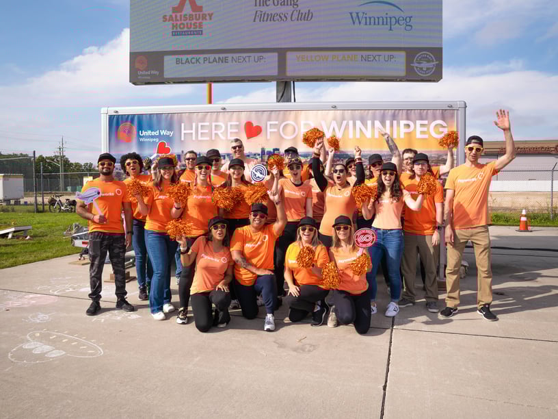 Un groupe de personnes qui portent des chandails orange avec le logo de Payworks agitent des pompons devant une pancarte sur laquelle on peut lire « Here for Winnipeg ».