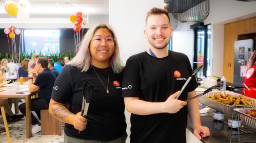 -	Deux membres du personnel manient des pinces de service devant des plateaux de déjeuner dans une cafétéria bondée et décorée de ballons..