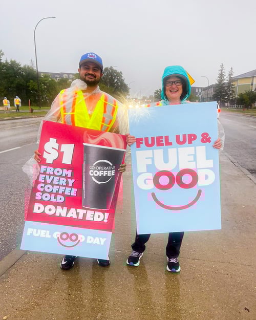 Two Payworks staff standing on a sidewalk in the rain. They’re wearing ponchos and holding up Fuel Up, Fuel Good signs. 