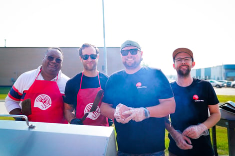 Four Payworks staff in United Way clothing in front of a barbecue. 