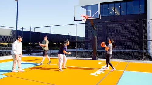 -	Quatre personnes durant une partie de basketball extérieur sur un terrain peint orange et bleu sarcelle, avec le logo de Payworks au sol.