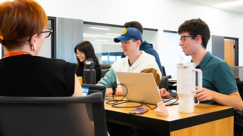 A group of five developers gathered around a table, chatting and working on laptops.