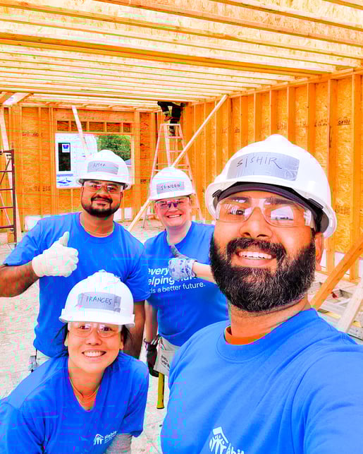 Quatre employés de Payworks portent des casques de sécurité arborant leur nom sur un morceau de ruban adhésif collé à l’avant et sourient à l’objectif sur un chantier d’Habitat pour l’humanité. 