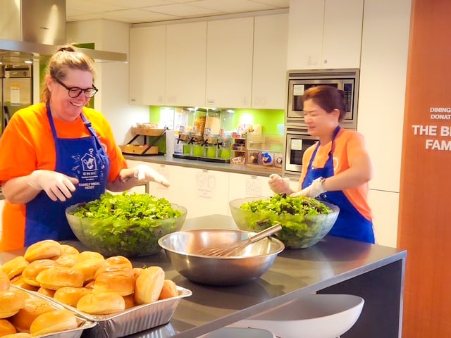 Two people wearing orange Payworks shirts and blue aprons preparing salad in a commercial kitchen.