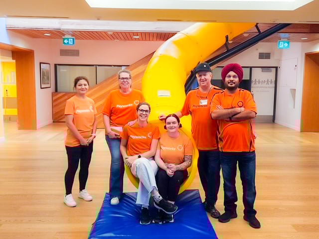 Six people wearing orange Payworks shirts sit or stand at the base of a big, yellow indoor tube slide.