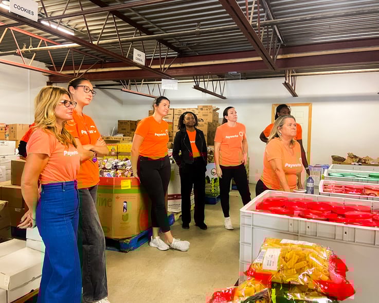 Un groupe de personnes en chandail orange Payworks écoute une personne hors champ dans un entrepôt rempli de dons alimentaires.