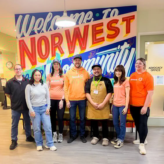 A group of people, several wearing orange Payworks shirts, standing in front of a colourful mural that reads “Welcome to NorWest.”