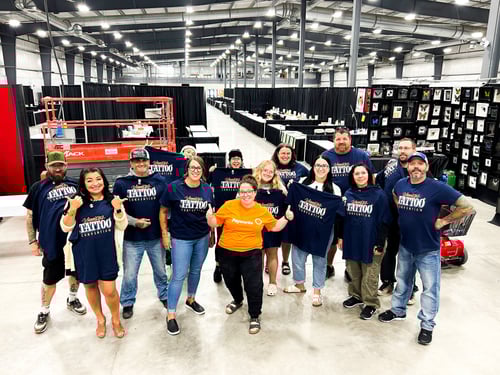 Un employé de Payworks avec un grand groupe de bénévoles portant des chandails de la Winnipeg Tattoo Convention dans une salle de congrès remplie de kiosques..