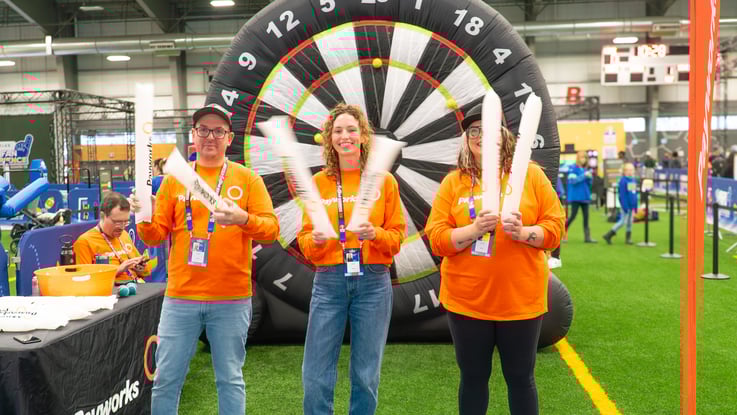 Trois personnes en chandail orange Payworks tapent des applaudisseurs gonflables sur le terrain d’un stade couvert, devant une cible de fléchettes géante gonflable.