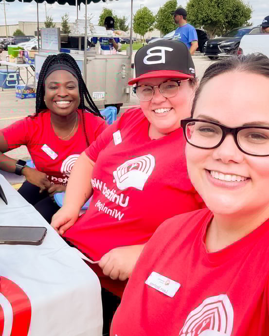 Three United Way Sponsored Campaign Associates smiling at a table in a parking lot on a sunny day.