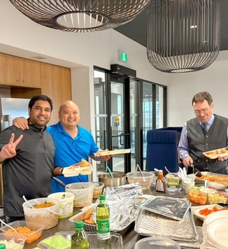 Three people serving themselves food and smiling for the camera behind a large buffet of homemade dishes spread out on an office kitchen counter.