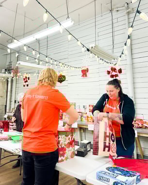 Two people wrapping gifts under string lights and wearing orange Payworks shirts. The words “pay it forward” are visible on the back of one.