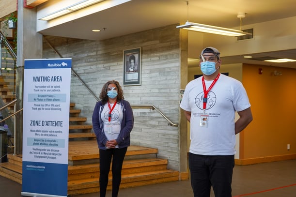 Deux personnes portant un masque et des protections pour les yeux se tiennent debout dans la salle d’attente du centre de vaccination pour les communautés autochtones urbaines de la Win Gardner Place.