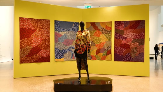 A large wall with four blanket art pieces handing on it. A mannequin stands on a platform in front wearing clothes by an Inuit designer.