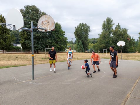 Trois adultes et trois enfants jouent au basket sur un terrain en extérieur.