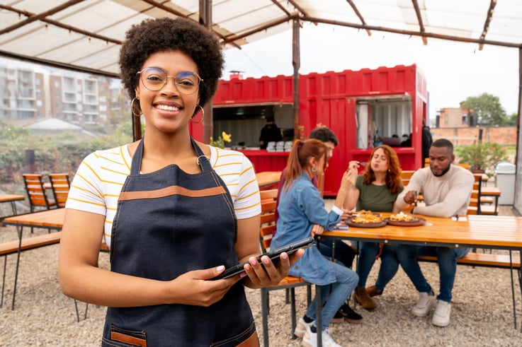 Une personne en tablier, debout sur la terrasse d’un restaurant, tenant une tablette.