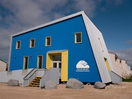 Le bâtiment de Polar Bears International : un bâtiment bleu avec un toit gris et une porte de couleur jaune vif. Le bâtiment est carré et son côté droit est incliné vers l’intérieur.