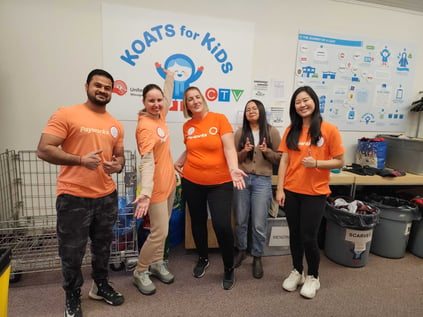 Cinq membres du personnel portant des t-shirts orange assortis se tenant devant une affiche Koats for Kids.
