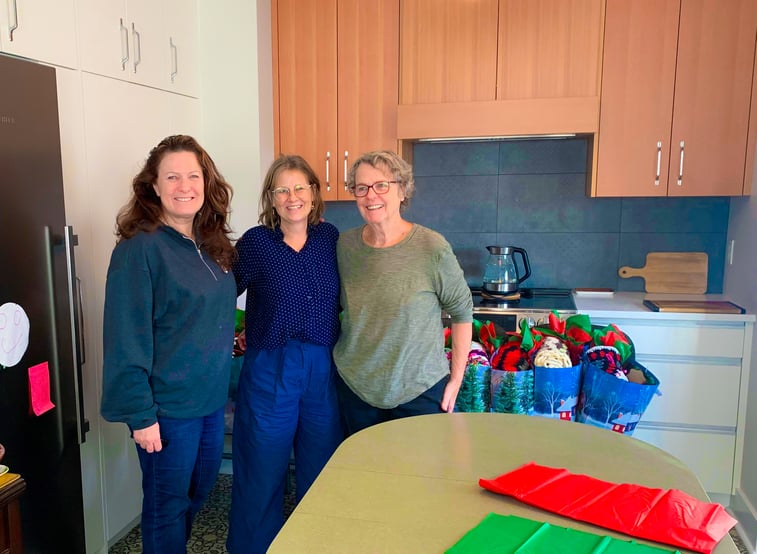 Trois personnes debout dans une cuisine résidentielle devant des sacs-cadeaux, avec du matériel d'emballage sur une table au premier plan.