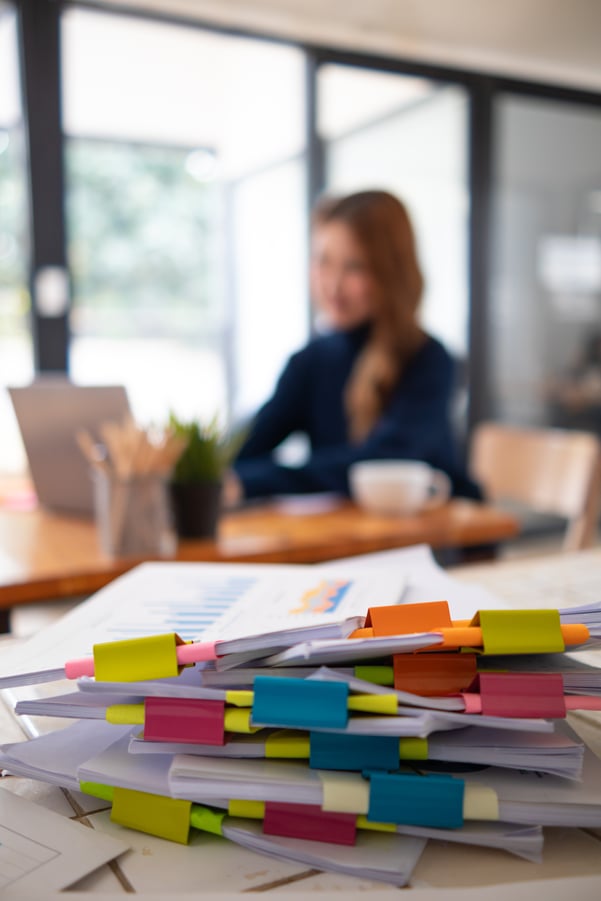 A stack of organized documents with a blurred woman at a computer in the background. 