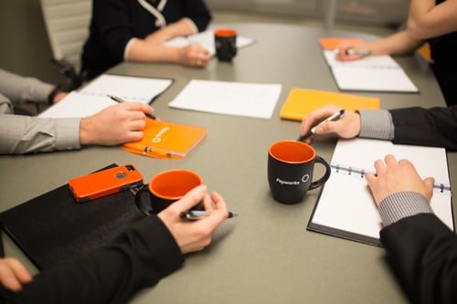 A table with Payworks mugs and notebooks on it and several people gathered around it, all of who are shown from the elbows down.