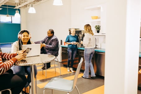 Five employees in a lunchroom. Two talk at the coffee machine, one works on a laptop, one works on a laptop with headphones on, and the other is on a tablet. 