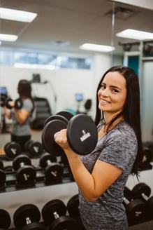 An individual in a gym, lifting a five pound weight and looking at the camera.