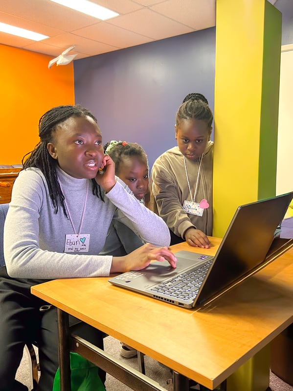 An instructor and two students sit side by side, looking at a laptop screen.