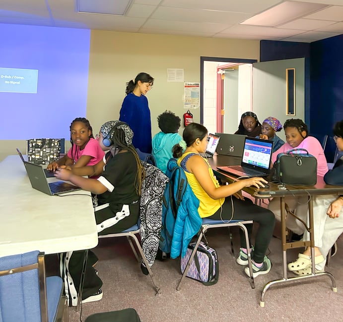 Two long tables full of students using laptops are overseen by an instructor.