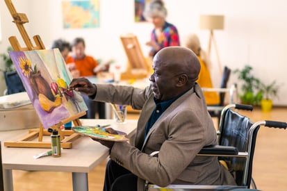 An art classroom. One individual is finishing a painting of a vase with a sunflower in it. They are sitting down in front of the canvas holding a palette and a paintbrush. 